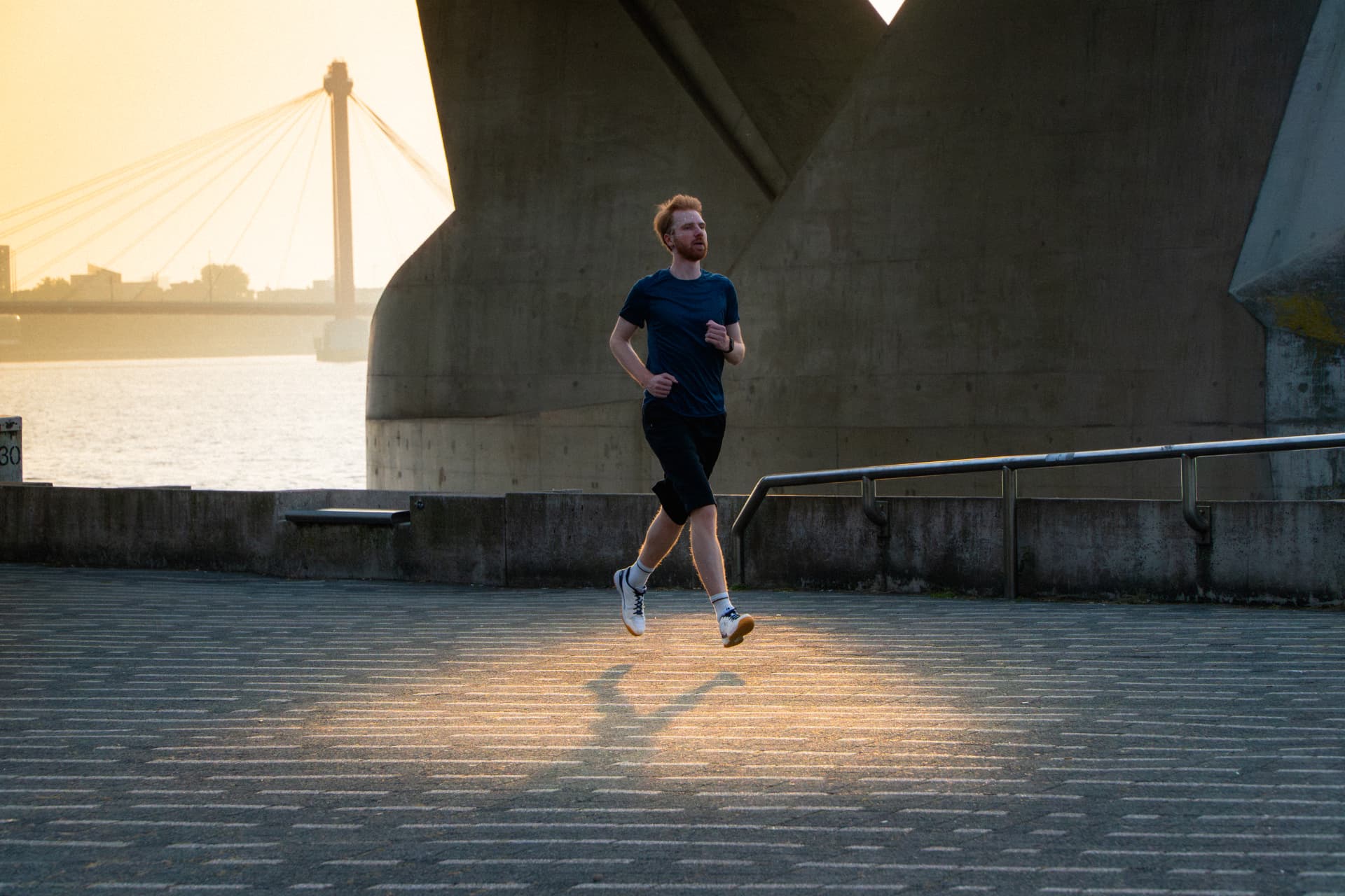 Runner jogging along a riverside under a concrete bridge at sunset