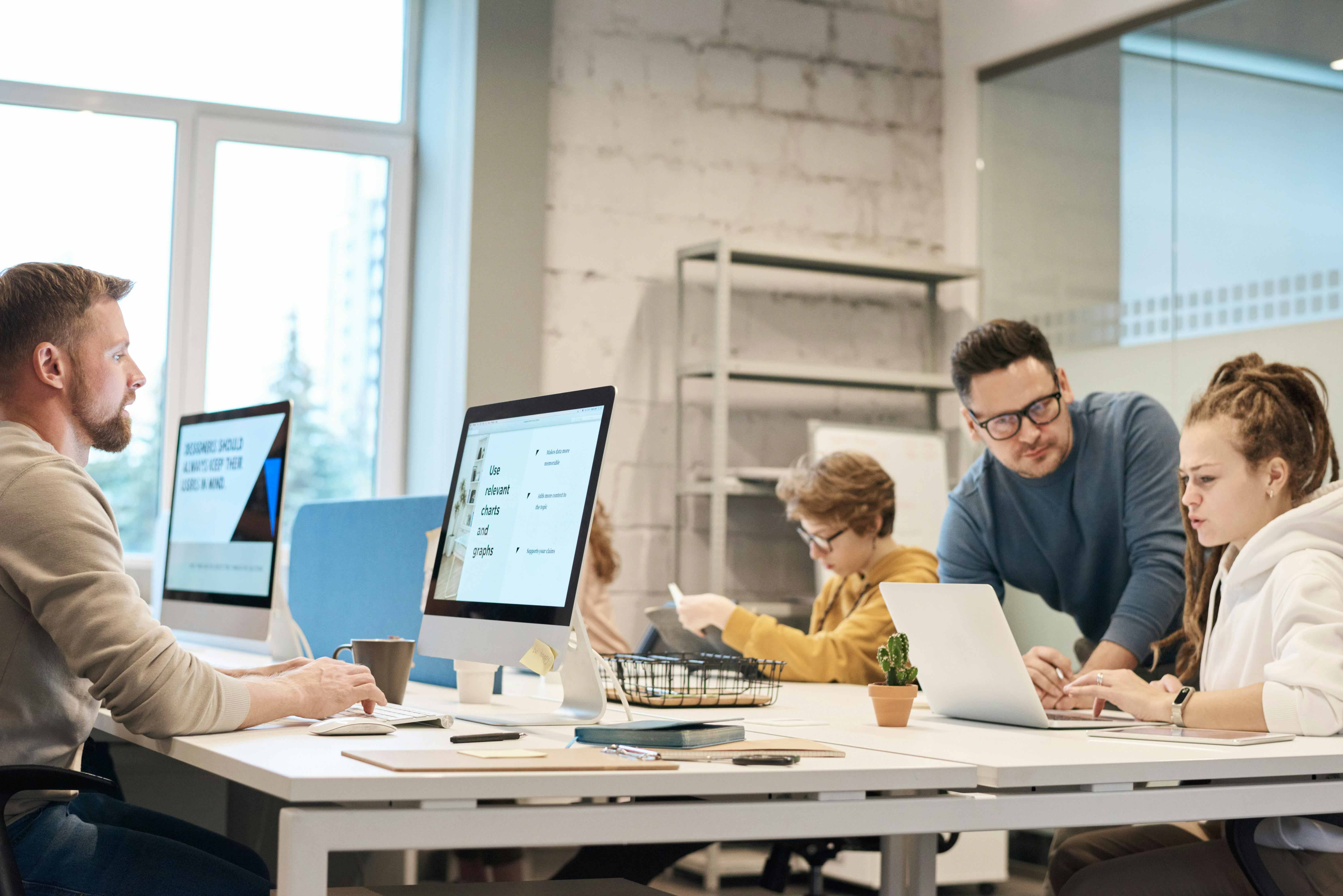 Four people working in the office in front of the computer screens. A woman and a man discuss something while looking at the computer screen together in the bottom right corner of the image.